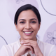Headshot portrait of a smiling South Asian mother with well-manicured hands, set against a soft lavender and white background