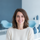 A smiling French woman in her late 20s, wearing a light top, against a blurred background with slate blue and white tones.